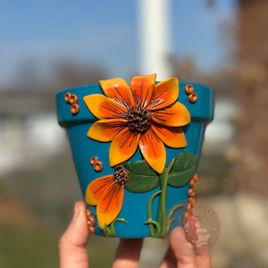 Pode incluir: Um vaso de flores de cerâmica azul-petróleo com um design de girassol em relevo. Os girassóis têm pétalas laranja e centros castanhos. Folhas e caules verdes também fazem parte do design, com pequenas contas castanhas.