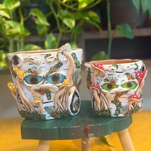May include: Two cylindrical ceramic planters with a white birch bark texture, featuring sculpted 3D mushrooms and large, detailed eyes. The larger planter has blue eyes and yellow mushrooms, while the smaller one has green eyes and red mushrooms.