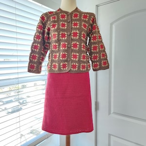 May include: A brown and red crocheted cardigan with a matching red crocheted skirt. The cardigan has a button closure and a square pattern.