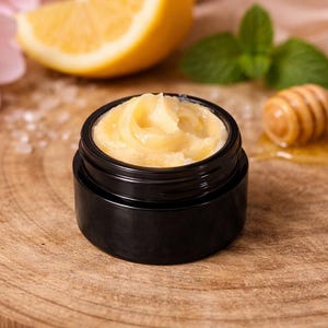 Könnte beinhalten: Ein kleines schwarzes Glas mit hellgelber Creme steht auf einer Holzoberfläche. Eine Zitronenscheibe, rosa Blüten und ein hölzerner Honiglöffel mit Honig befinden sich im Hintergrund. Die Creme hat eine glatte, gewirbelte Textur.