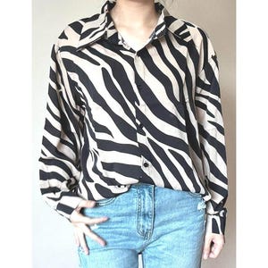 Pode incluir: Camisa de manga comprida com padrão de zebra em preto e bege. A camisa tem gola e punhos clássicos. A modelo está usando jeans azuis. A camisa é abotoada na frente.