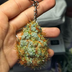 Może przedstawiać: Ręcznie robiony brelok do kluczy z zielono-szarą, teksturowaną zawieszką w kształcie pąka z pomarańczowymi i srebrnymi akcentami. Zawieszka jest przymocowana do srebrnego łańcuszka i kółka na klucze. Brelok jest trzymany w dłoni.