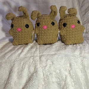 May include: Three brown crocheted bunny plushies with pink noses and black eyes. The plushies are sitting on a white fluffy surface.