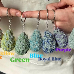 May include: A collection of handmade, fluffy keychains in various colors, including pink, yellow, green, blue, purple, grey, and white. Each keychain features a silver-tone keyring and chain. The keychains are displayed against a neutral background.