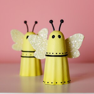 May include: Two yellow bee-shaped figures crafted from egg carton segments. Each figure features two black antennae, two black eyes, a black horizontal band with dots, and shimmering, glittery white wings attached to the sides of the body.