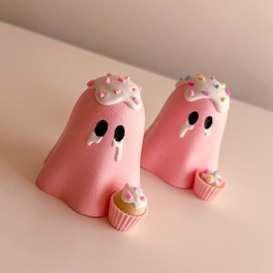 May include: Two pink ghost figurines with black oval eyes and white icing drips on their heads topped with colorful sprinkles. Each ghost is positioned next to a small, light-colored cupcake in a pink pleated paper liner with white icing.