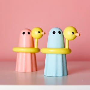 May include: Two small wooden figurines shaped like ghosts, one in pastel pink and one in light blue. Each ghost wears a bright yellow ring around its middle and has a yellow bird-like figure attached to its side with a small orange beak.
