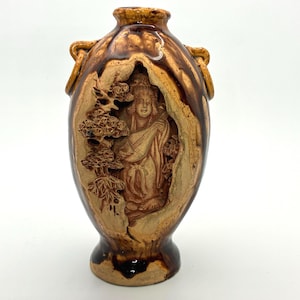 May include: A brown ceramic vase with a carved relief of a figure playing a stringed instrument, set within a landscape of trees. The vase has a dark brown base and rim, with two small handles on the sides.