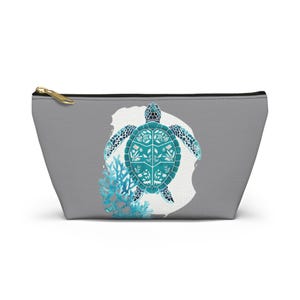 Könnte beinhalten: Graue Reißverschlusstasche mit einem Design einer türkisfarbenen Meeresschildkröte mit floralen Mustern auf ihrem Panzer. Das Design umfasst blaue Korallen und einen weißen Hintergrund. Die Tasche hat einen goldenen Reißverschluss.