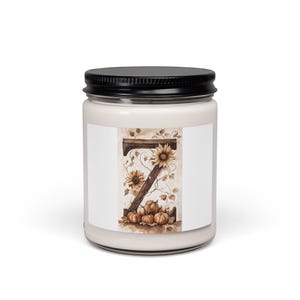 May include: A glass candle jar with a black lid containing white wax. The label features a large letter Z formed from wood, decorated with brown sunflowers, swirling vines, and a cluster of small orange pumpkins at the base of the letter.