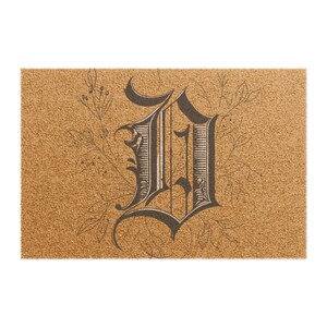 May include: A rectangular tan coir doormat featuring a large, dark brown, ornate gothic-style letter D centered on the surface. The letter is surrounded by delicate, thin-lined botanical illustrations of leaves and stems in a dark brown ink.