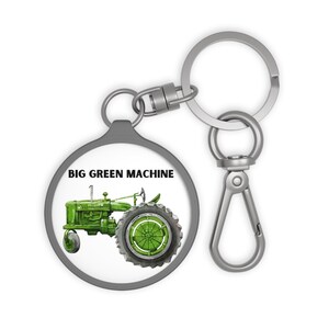 以下が含まれることがあります： グレーの縁とシルバーの留め金が付いた丸いキーホルダー。キーホルダーには、黒い文字で「BIG GREEN MACHINE」と書かれた緑色のトラクターのイラストが描かれています。トラクターは大きな後輪と小さな前輪を持っています。