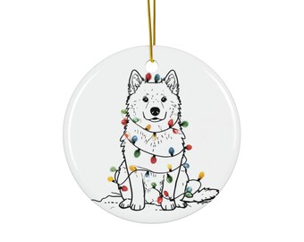 May include: A round, white ceramic ornament featuring a black and white illustration of a dog wrapped in colorful Christmas lights. The ornament has a gold string for hanging, perfect for holiday decor.