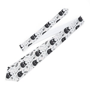 May include: A white necktie with a repeating pattern of black cats and grey leaves. The cats have pink noses and white eyes.