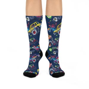 May include: A pair of knee-high socks with a repeating pattern of colorful monster trucks on a dark blue background. The socks have black heels and toes. The monster trucks are yellow, red, green, and purple.