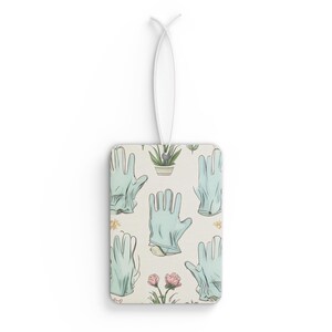 May include: Rectangular air freshener with a white string. The air freshener features a pattern of light blue gardening gloves, potted plants, and pink flowers on a cream background. The design is repeated across the surface.