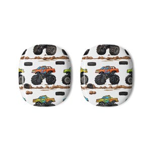 May include: Two white, oval-shaped buttons with a repeating pattern of colorful monster trucks, tires, and dirt. The trucks are orange, green, and yellow with black tires. The background is white.
