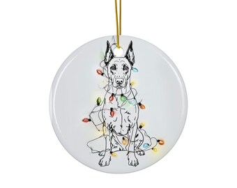Peut inclure: Ornement de Noël en céramique blanche avec une illustration en noir et blanc d'un chien orné de guirlandes lumineuses colorées. L'ornement est suspendu à une ficelle dorée.