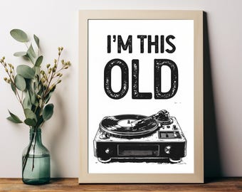 May include: A framed print with the text "I'M THIS OLD" above a black and white illustration of a record player. The frame is light-colored, and the print is displayed on a wooden surface with a vase of greenery.