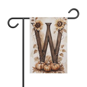 Peut inclure: Un drapeau de jardin rectangulaire suspendu à un support en métal noir. Le drapeau présente une grande lettre W à la texture de bois brun foncé, ornée de deux tournesols jaunes en haut et de trois citrouilles orange avec des feuilles brunes.