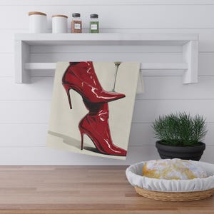 May include: A red high-heeled boot illustration on a cream-colored towel. The boots are glossy and have pointed toes. A martini glass is next to the boots. The towel is hanging on a white shelf above a wooden countertop.