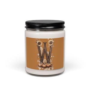 May include: A white candle in a glass jar with a black lid. The label features a large, dark brown letter W decorated with sunflowers and small pumpkins against a solid orange background. The design includes detailed illustrations of autumn foliage.