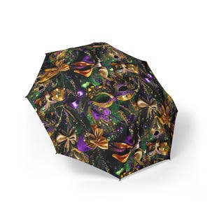 May include: A black umbrella with a festive pattern of gold and purple bows, masks, and beads. The design is set against a dark green background, creating a celebratory and eye-catching accessory.