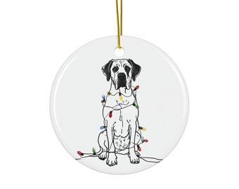 Könnte beinhalten: Runder, weißer Keramik-Weihnachtsschmuck mit einer schwarz-weißen Strichzeichnung eines Hundes, der mit bunten Lichterketten geschmückt ist. Der Anhänger hat einen goldenen Aufhänger.