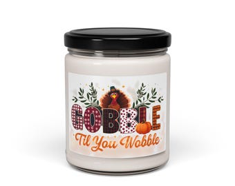 May include: A white candle jar with a black lid. The label features the text "GOBBLE Til You Wobble" in a colorful, patterned font, along with a turkey illustration and a pumpkin. The design is festive and suitable for Thanksgiving.