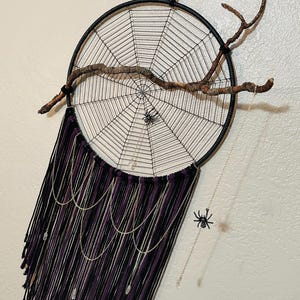 Könnte beinhalten: Ein schwarzer und weißer Traumfänger mit einem Spinnennetz-Design und einem Zweig, der von oben hängt. Der Traumfänger hat violette und schwarze Fäden, die von unten hängen, mit silbernen Ketten und Kristallen.