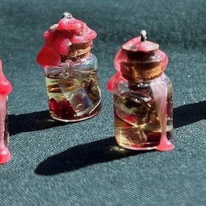 Puede incluir: Cuatro pequeñas botellas de vidrio con tapones de corcho, llenas de un líquido dorado y elementos botánicos. Cada botella está coronada con una mancha de cera roja brillante, parte de la cual gotea por los lados. Sobre una superficie texturizada verde oscuro.