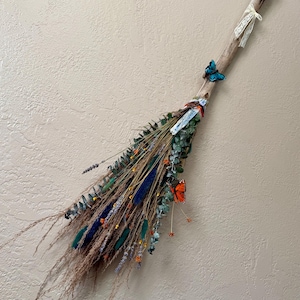 Puede incluir: Una escoba decorativa con un mango de madera adornado con flores secas, plumas y mariposas artificiales en tonos azules, naranjas y verdes. La escoba está adornada con una cinta de encaje y cristales.
