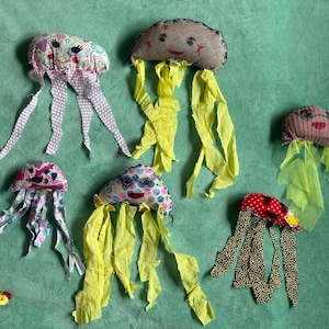 Puede incluir: Juguetes de medusas de tela hechos a mano en varios colores y patrones. Las medusas tienen caras sonrientes y largos tentáculos que fluyen. Los juguetes están hechos de diferentes telas, incluyendo lunares y estampados florales. El fondo es de color verde liso.