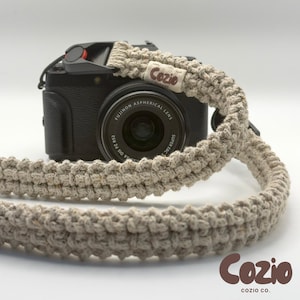 May include: A black digital camera with a Fujinon Aspherical lens attached to a thick, beige, macrame-style woven camera strap. The strap features a small, rectangular fabric tag with the word Cozio printed in brown text on the front.