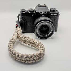 May include: A black Fujifilm X-T100 digital camera with a Fujinon Aspherical Lens 15-45mm attached. A thick, beige, hand-knotted macrame wrist strap is connected to the side of the camera body, featuring a small fabric tag near the attachment point.