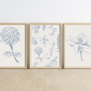Puede incluir: Tres impresiones enmarcadas con ilustraciones florales azules. La impresión de la izquierda presenta una sola flor azul sobre un fondo blanco. La impresión central presenta un patrón repetitivo de flores y hojas azules sobre un fondo blanco. La impresión de la derecha presenta una sola flor azul sobre un fondo blanco.