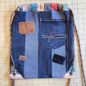 Op de afbeelding: Een blauwe denim trekkoordtas met een patchwork ontwerp. De tas heeft een groot vak aan de voorkant met de tekst "American Eagle" en een kleiner vak met de tekst "Lee".