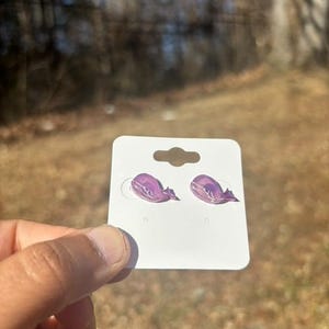 Puede incluir: Un par de pendientes morados con forma de ballena en una tarjeta blanca para pendientes. Los pendientes son de color morado claro con acentos morados más oscuros. La tarjeta tiene un agujero en la parte superior para colgar. El fondo está borroso, con tonos marrones y verdes.