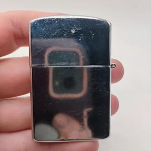 May include: A polished, silver-coloured lighter with a rectangular form. It features a hinged lid and a small, circular attachment. The lighter's surface reflects the surrounding environment, showing a distorted image.