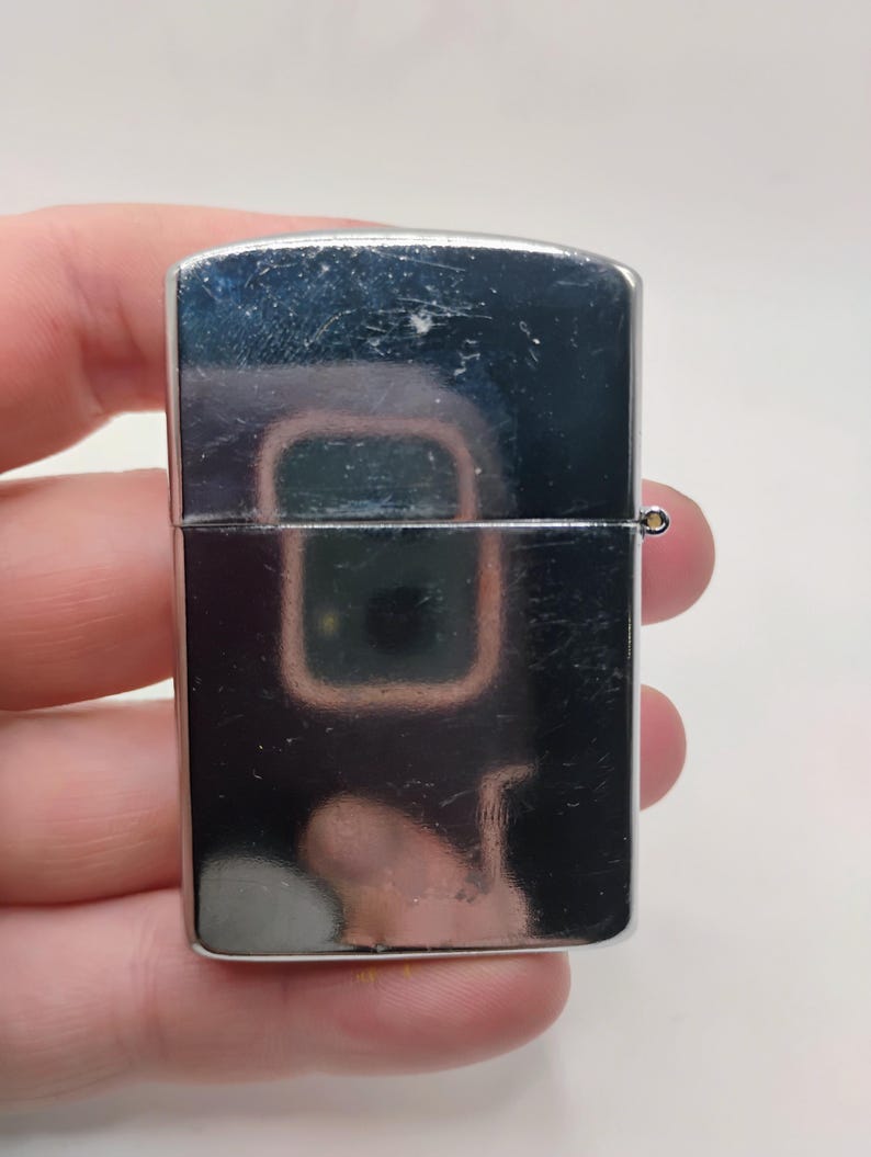 May include: A polished, silver-coloured lighter with a rectangular form. It features a hinged lid and a small, circular attachment. The lighter's surface reflects the surrounding environment, showing a distorted image.