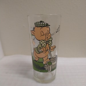 Op de afbeelding: Een helder glas met een cartoonafbeelding van een varken dat een hoed en een strik draagt, en met een grasmaaier het gras maait. Het varken blaast een melodie uit zijn neus.
