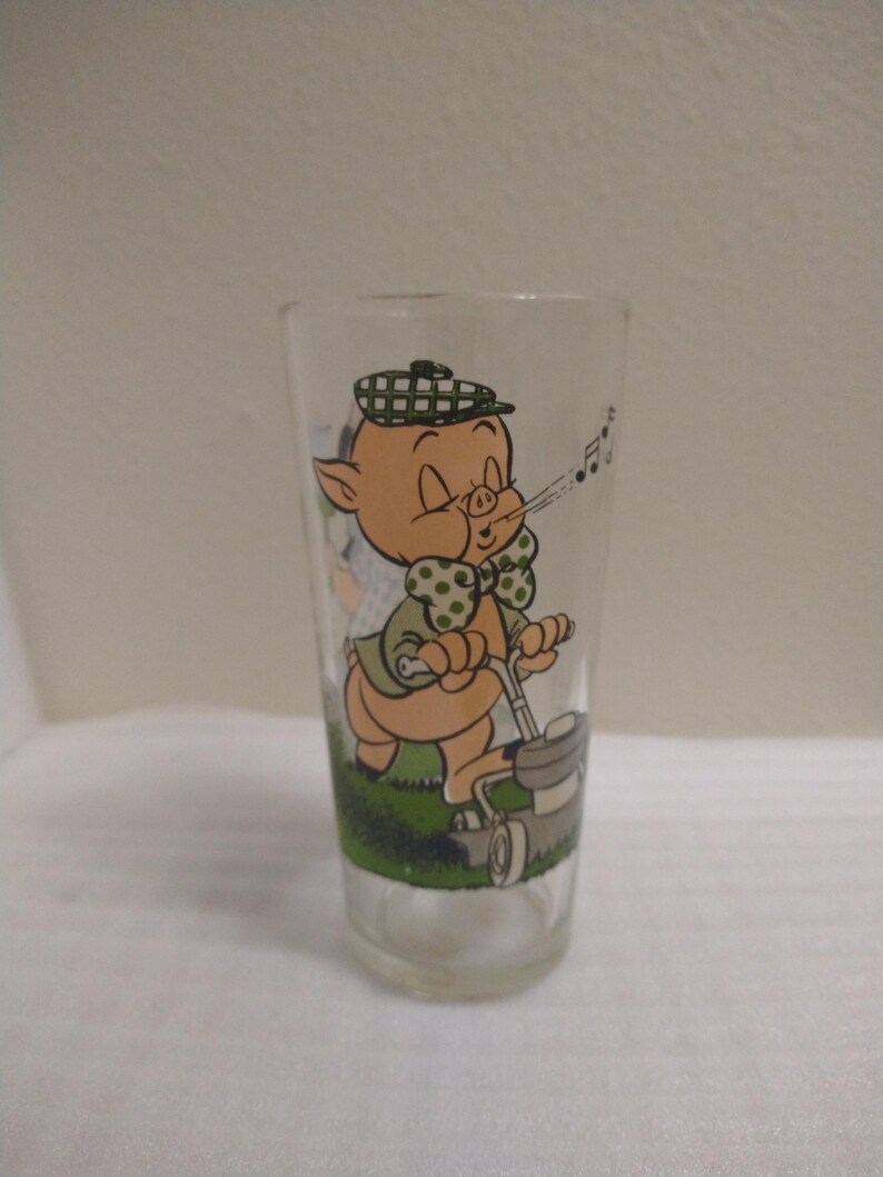 Op de afbeelding: Een helder glas met een cartoonafbeelding van een varken dat een hoed en een strik draagt, en met een grasmaaier het gras maait. Het varken blaast een melodie uit zijn neus.