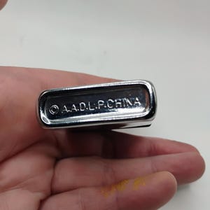May include: A polished, silver-coloured lighter with a rectangular shape and rounded corners. The text "&copy; A.A.D.L.P.CHINA" is engraved on the base. The lighter is held in a hand.