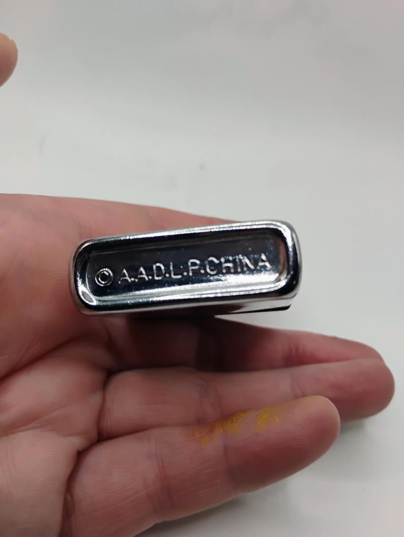 May include: A polished, silver-coloured lighter with a rectangular shape and rounded corners. The text "&copy; A.A.D.L.P.CHINA" is engraved on the base. The lighter is held in a hand.