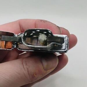 May include: Close-up of a silver metal lighter with a rectangular shape. The lighter's interior reveals a wick and other components. The lighter is held in a hand against a white background.