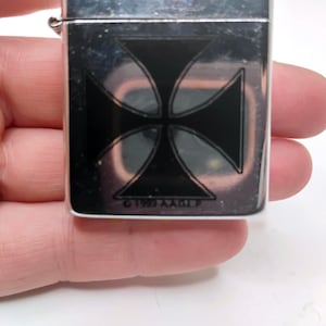 May include: Silver metal lighter featuring a black Maltese cross design. The rectangular lighter has a hinged lid. The text "&copy;1993 A.A.O.L.P" is printed at the bottom of the cross.