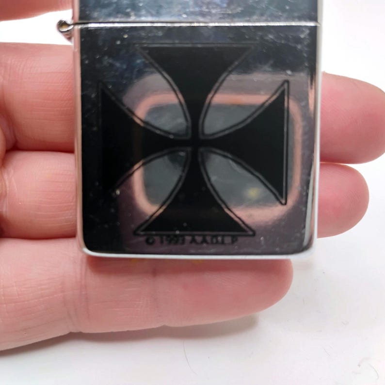 May include: Silver metal lighter featuring a black Maltese cross design. The rectangular lighter has a hinged lid. The text "&copy;1993 A.A.O.L.P" is printed at the bottom of the cross.