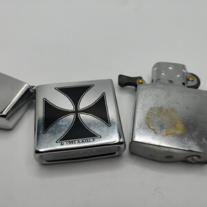 May include: Three silver lighters are shown. One is open, displaying a black and silver Maltese cross design. The other two lighters are closed. The lighters have a metallic finish and show signs of wear.