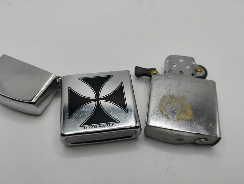May include: Three silver lighters are shown. One is open, displaying a black and silver Maltese cross design. The other two lighters are closed. The lighters have a metallic finish and show signs of wear.