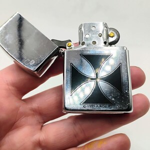 May include: A silver metal lighter featuring a black cross design. The lid is open, showing the inside. The text "&copy; 1993 A.A.O.L.P." is printed on the lighter.